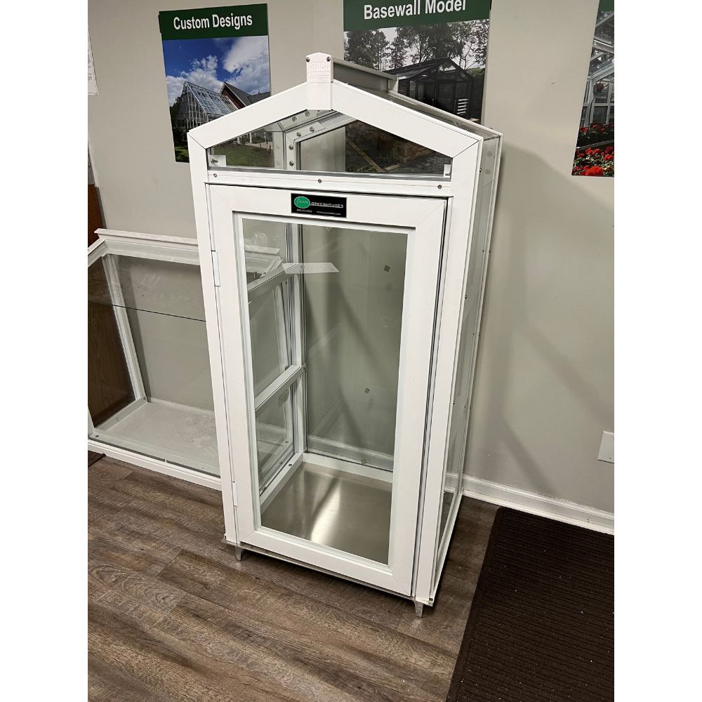 Front view of white Janco Garden Curio indoors with glass door and greenhouse-style peaked roof