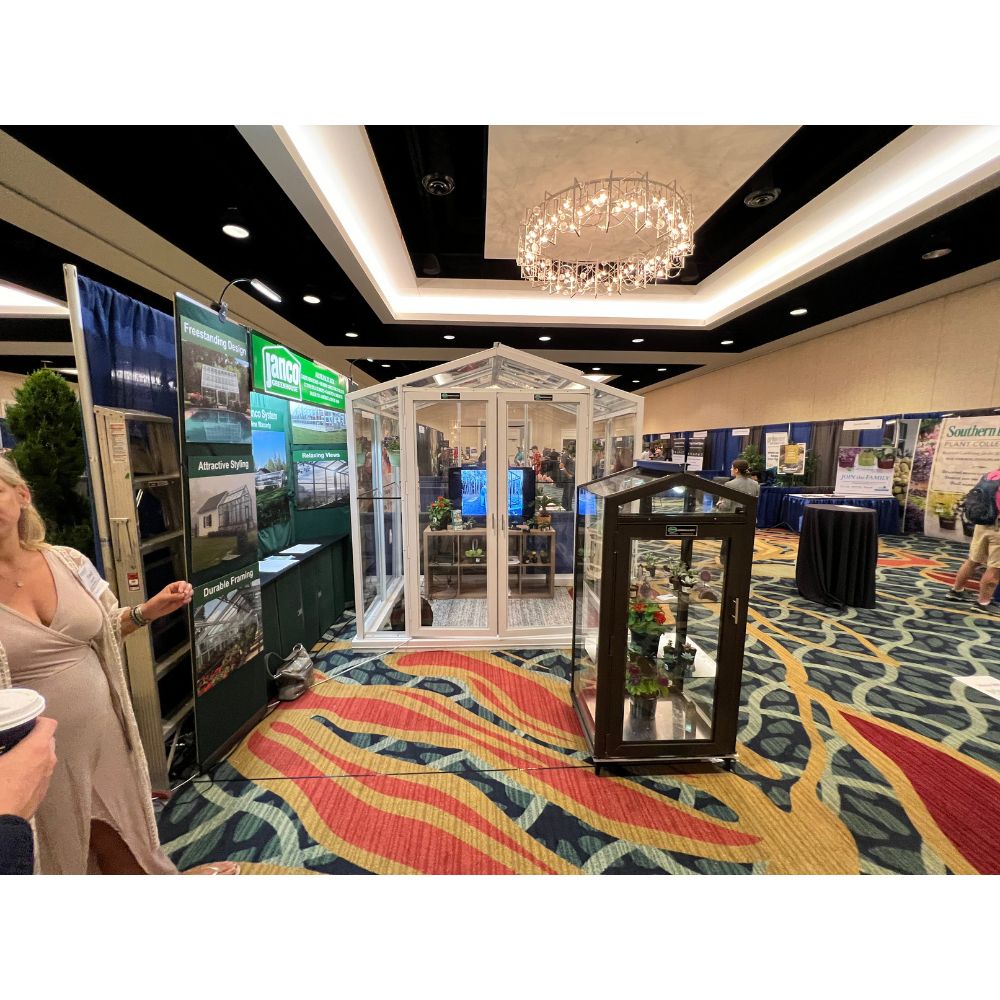 Janco Sago 5' x 8' white aluminum and glass greenhouse displayed at a trade show