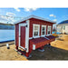 A red and white wooden chicken coop with three front windows, a door, and a front nesting box, photographed outdoors on a dirt lot under a blue sky.