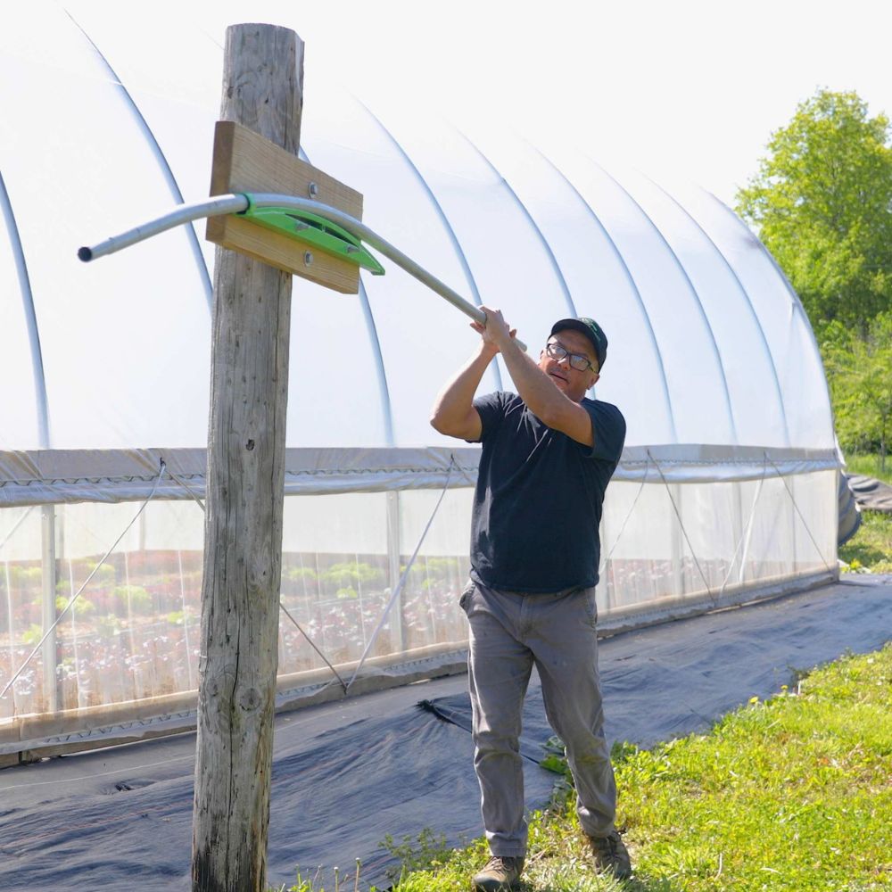 Person assembling a metal arch for the Bootstrap Farmer DIY Gothic Greenhouse beside a covered greenhouse structure